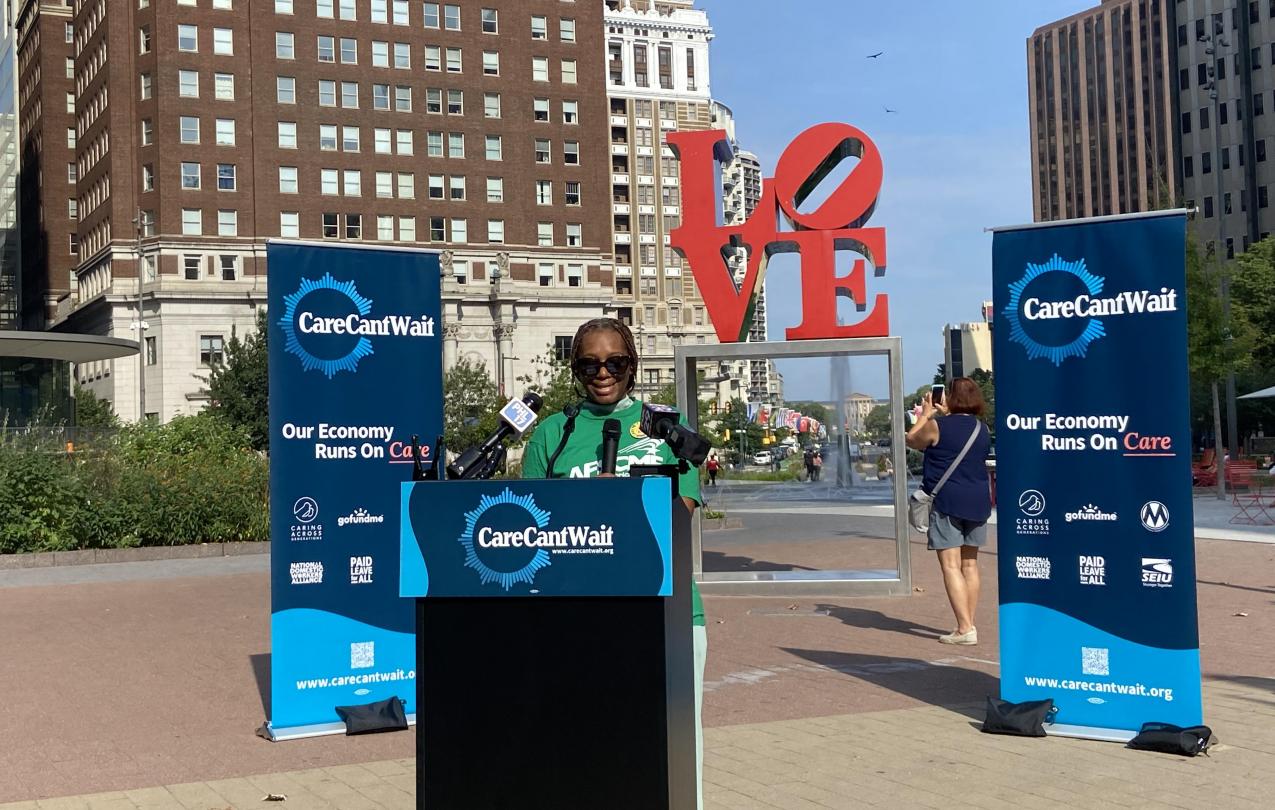 Woman in green AFSCME shirt speaking at podium in front of Care Cant Wait banners. 